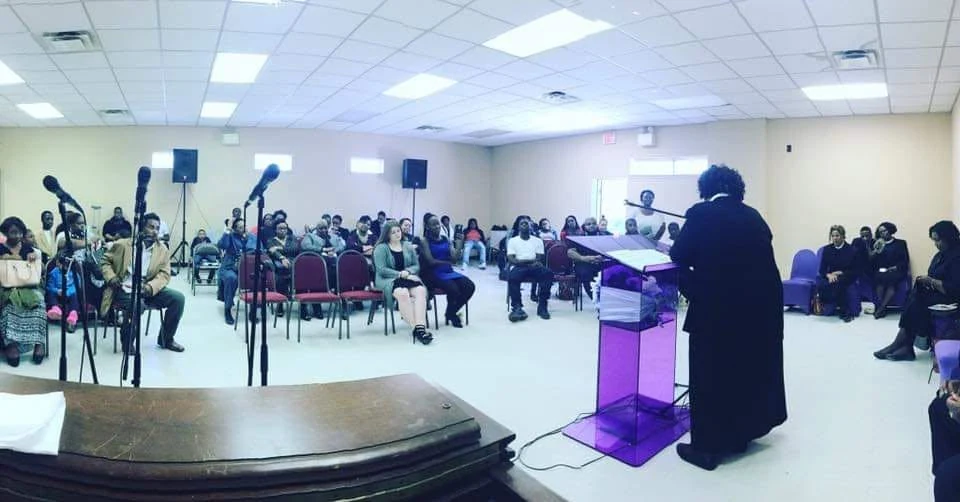 Overseer Betterson preaching from a purple acrylic pulpit to a full room
