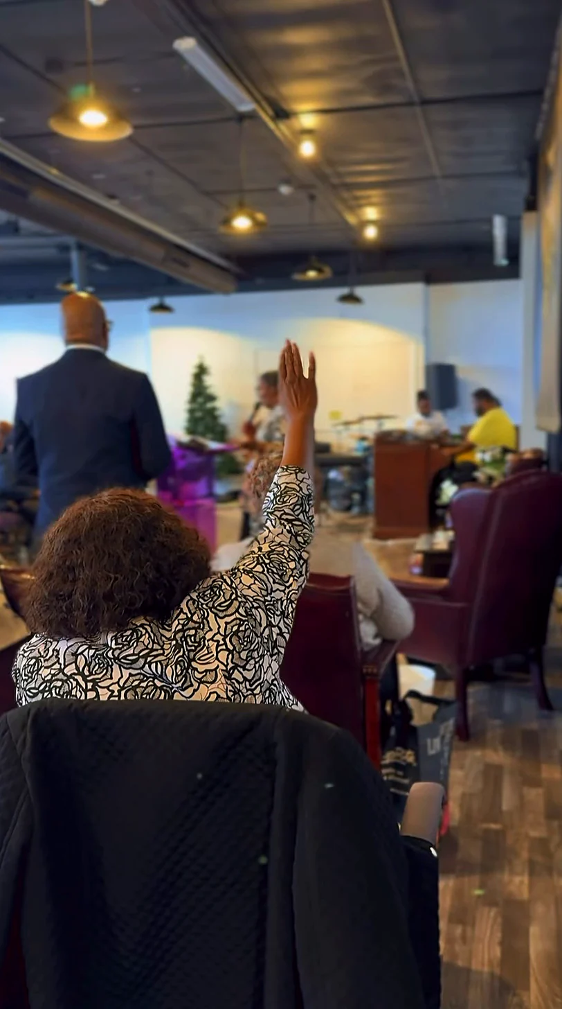 A worshiper lifts a hand in praise during Sunday service, the room full of purple chairs and the band up front